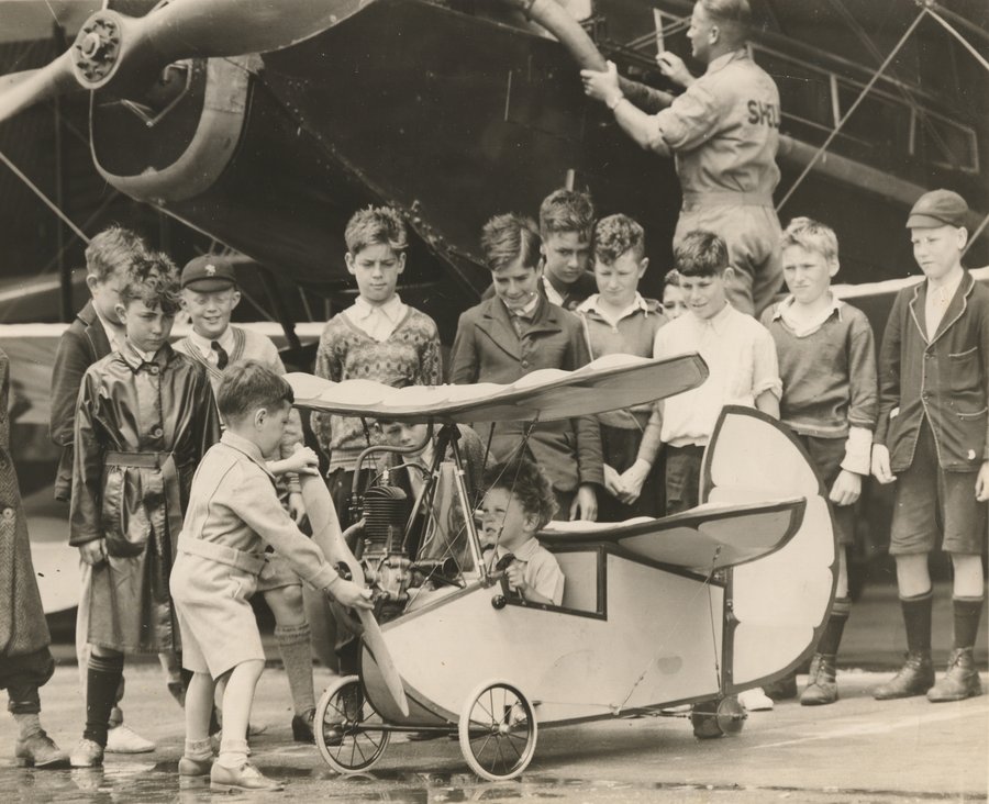 Howard Morris spinning the propeller of the Flying Flea with his brother Eric seated in the cockpit, watched by a crowd of boys, Essendon Aerodrome